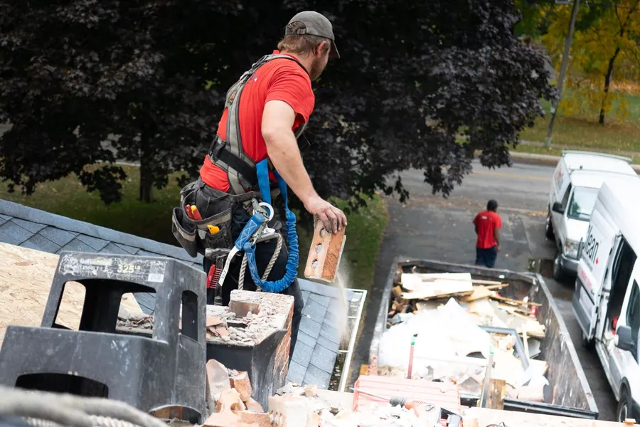 Worker in a safety harness lowering roofing material into a waste container below