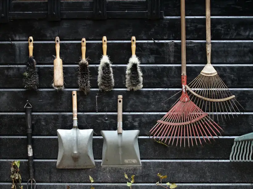 Garden tools and a rake hanging on the dark timber-clad wall of a garden outbuilding