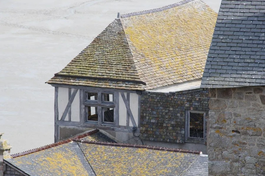Stone and timber house with gray and yellow shingled roofs, seen from above against a pale sky