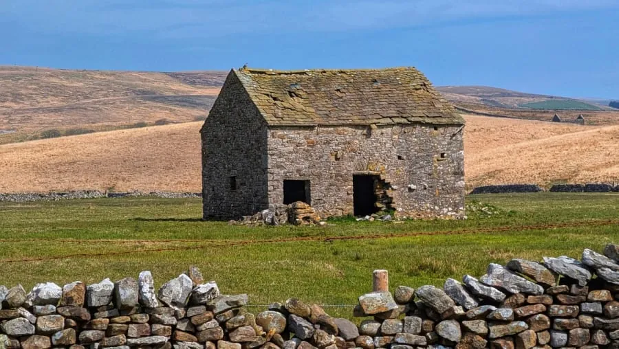 Small stone cottage in a grassy field, with dry hills in the background and a stone wall in front.