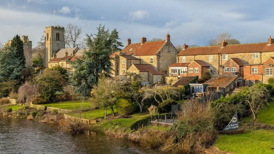 Riverside village with stone houses and a church tower beside a calm river under a cloudy sky