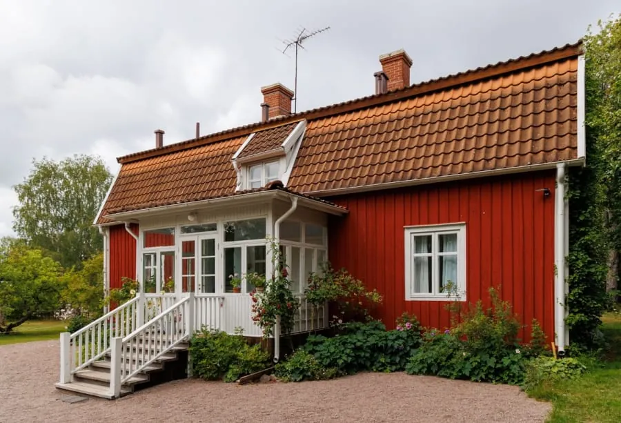 Red wooden house with white porch and orange tiled roof, surrounded by greenery