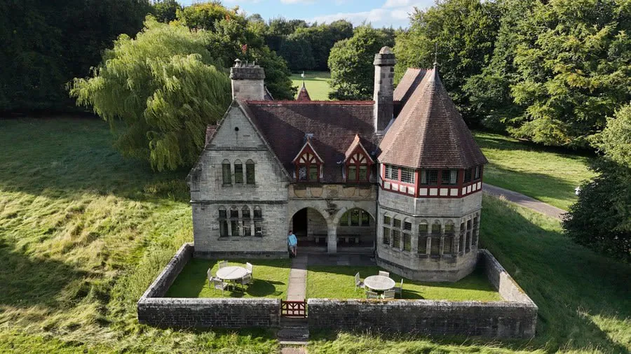 Miniature stone house with red roof and turret, surrounded by a grassy moat and trees.
