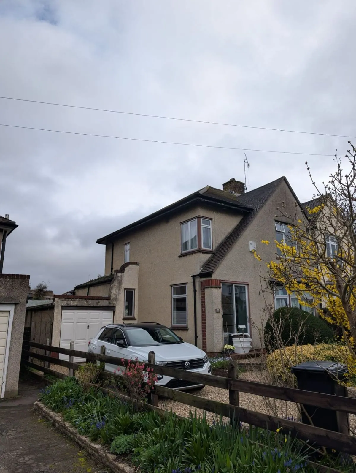 Two-story beige house with parked silver car, wooden fence, and cloudy sky