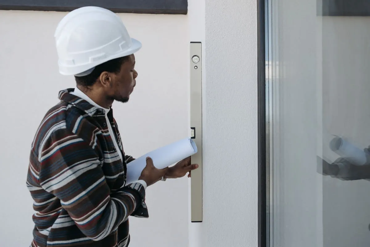 Worker in a white hard hat holding papers beside a glass door and wall-mounted access panel