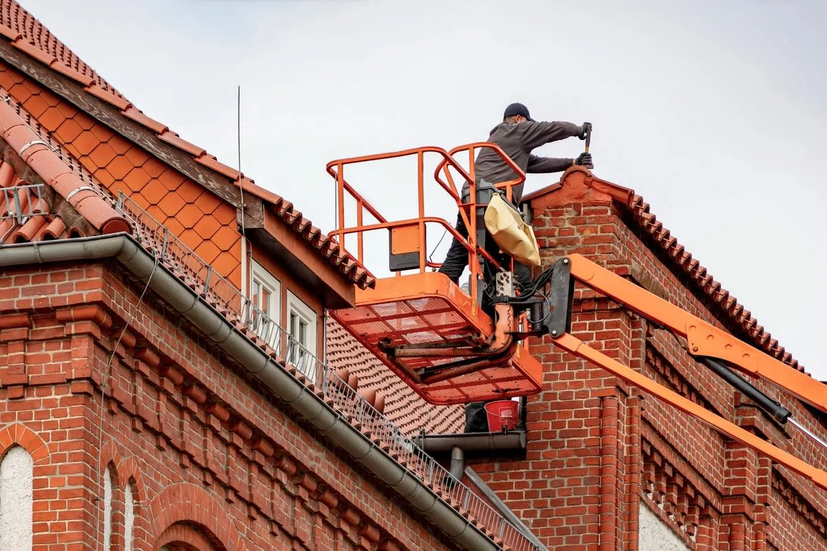 Worker on a roof using an orange lift to repair red brick and tiled building roof.