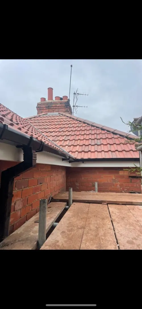 A view of a red-brick wall and tiled roof with a brick chimney and antenna against a cloudy sky.