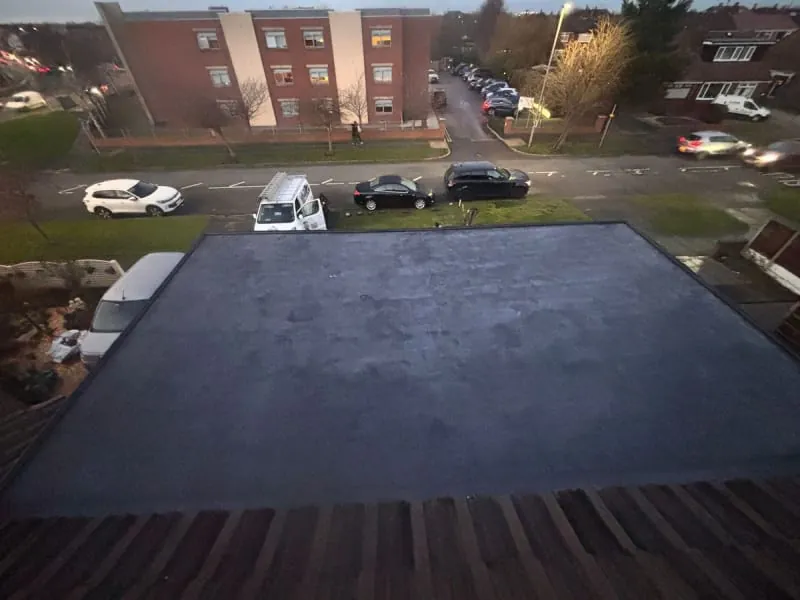 A high-angle view of a flat, dark gray rooftop above a residential street with parked cars and a brick building beyond.