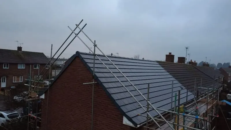 A residential house roof under repair with scaffolding and ladders set up against a grey, overcast sky.