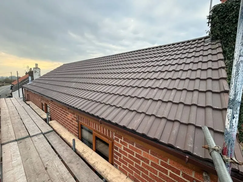 A side view of a residential brick house roof with new brown tiled roofing, seen from an adjacent scaffolding platform.