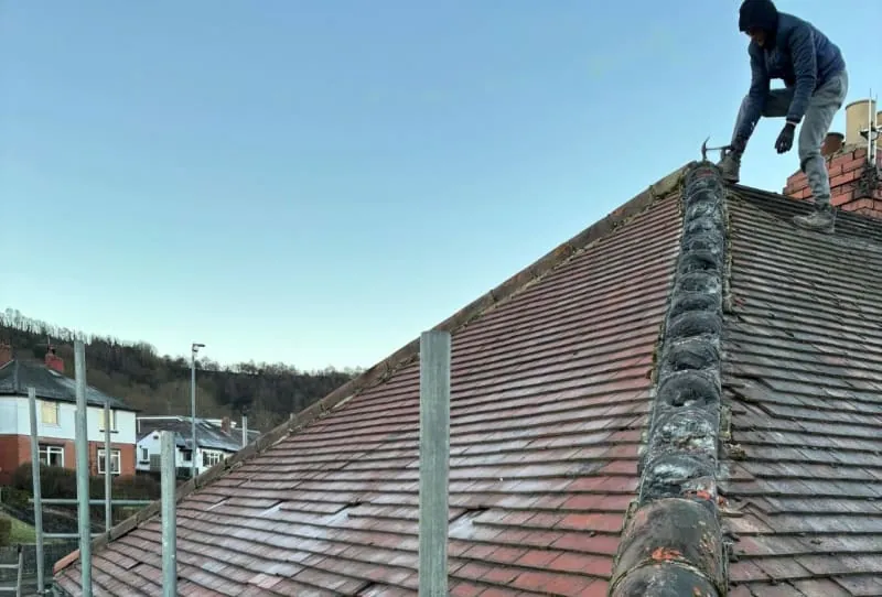 A person works on a tiled roof ridge, with metal scaffolding set up alongside the building against a clear sky.
