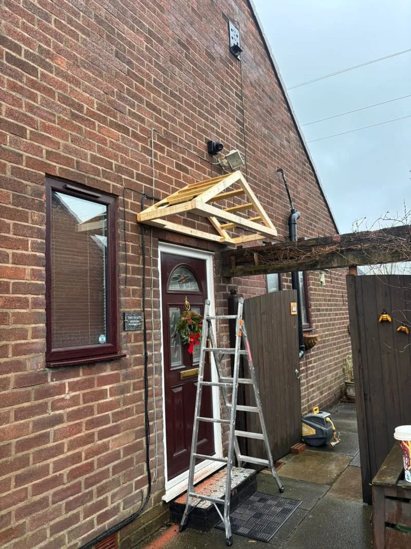 A wooden canopy frame is mounted above an exterior door, with an aluminum ladder standing in front of the doorway.