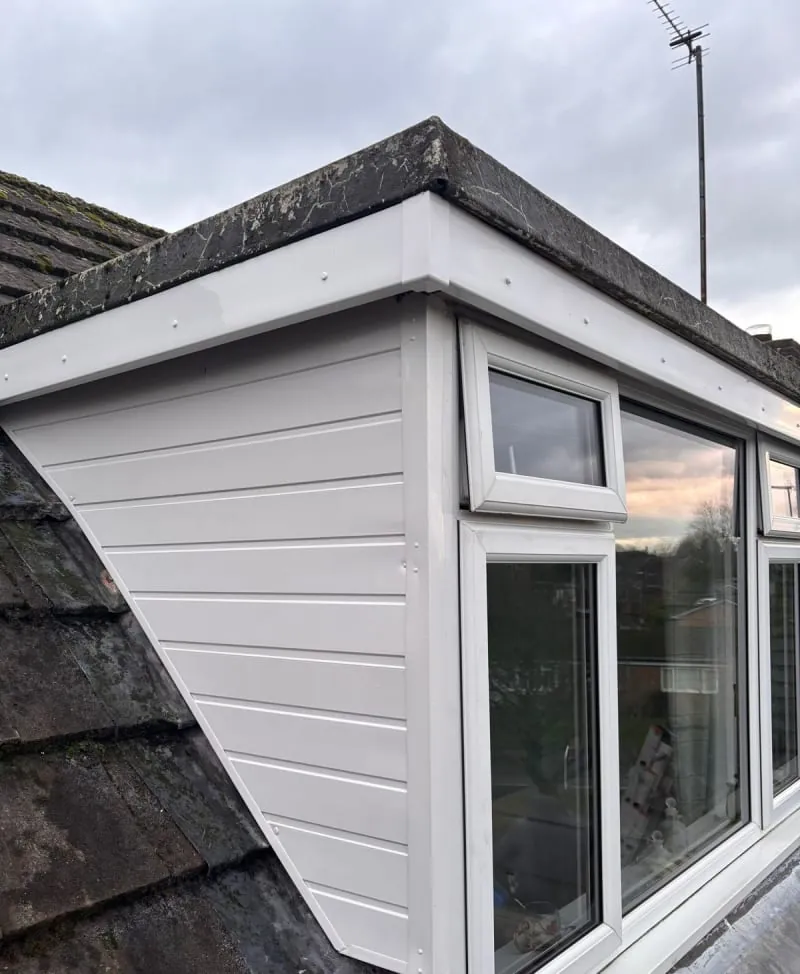A white dormer window with horizontal cladding and white trim, viewed from the side against a roof with dark tiles.