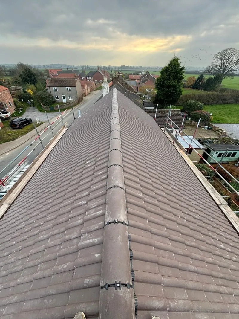 A high-angle view down the ridge of a brown tiled roof, overlooking a road, surrounding houses, and green fields at dusk.