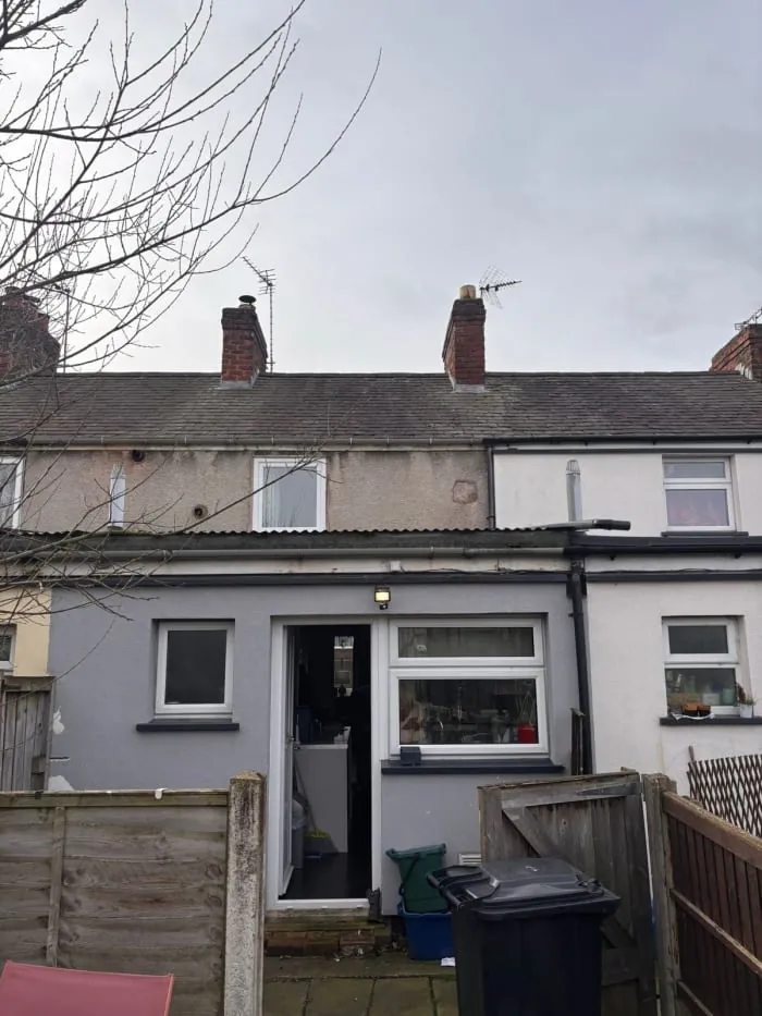 A grey, single-story rear house extension with a doorway and window, attached to a traditional two-story brick row house.