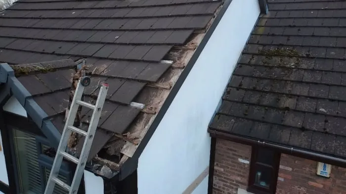 A ladder leans against a residential roof with missing tiles and exposed debris along the edge of a white wall.