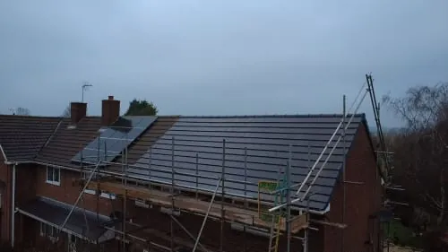 A residential brick house with a partially tiled roof, surrounded by scaffolding and featuring installed solar panels.