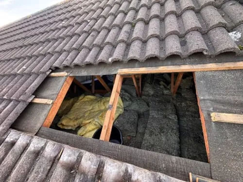 A section of roof tiles removed to reveal wooden rafters and yellow fiberglass insulation inside an attic.